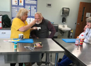 A woman in a gold t-shirt holds a tasting spoon up to a man with a beard and brown hoodie shirt, so he can taste what's on the spoon. a couple on the right are laughing.
