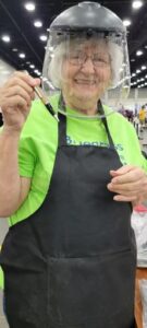 A woman wearing a safety shield, bright green t-shirt and black apron smiles at the camera while she holds up a wooden pen